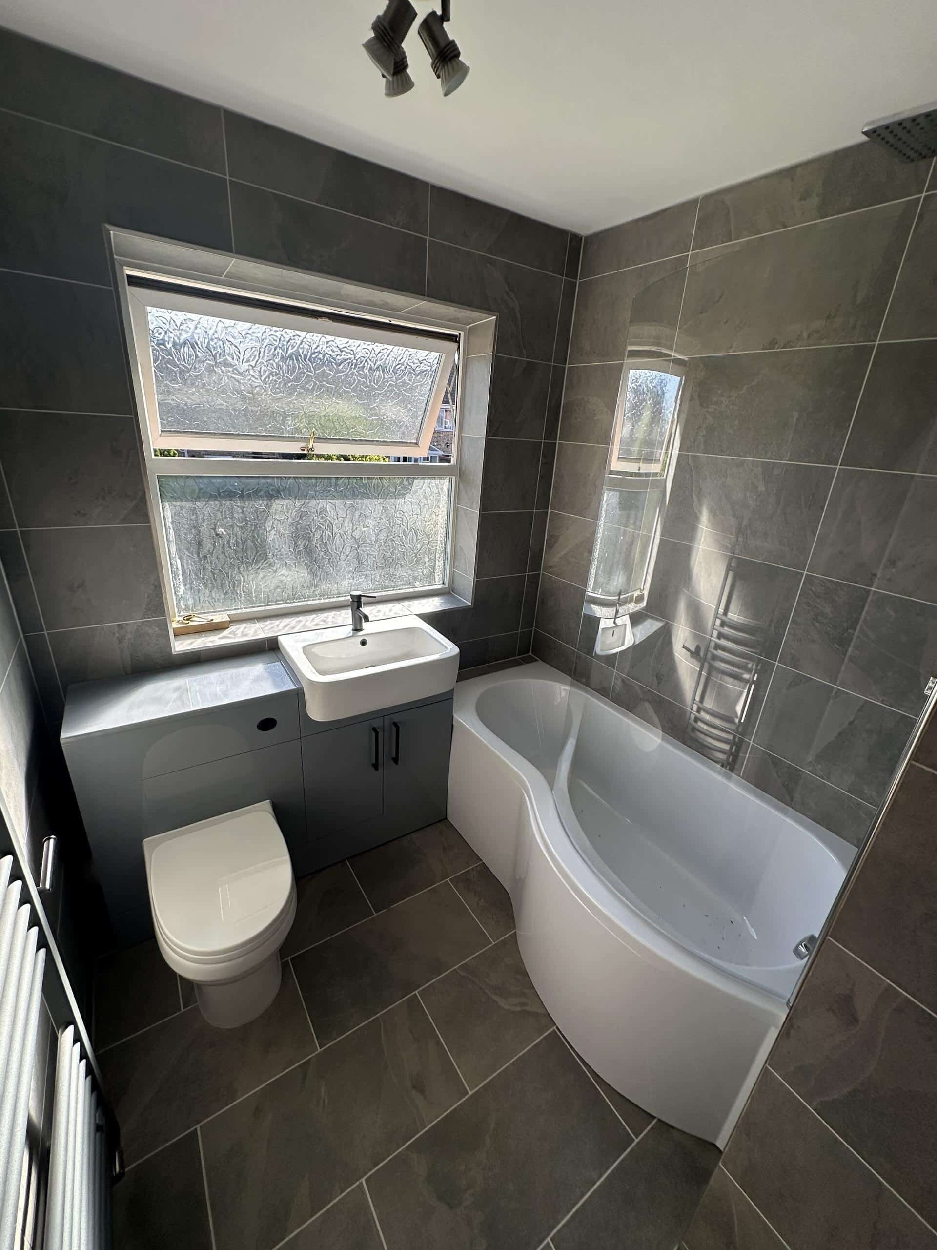 Bathroom Installations (1) Modern bathroom with a bathtub, sink, and toilet next to a frosted window and grey tiled walls.