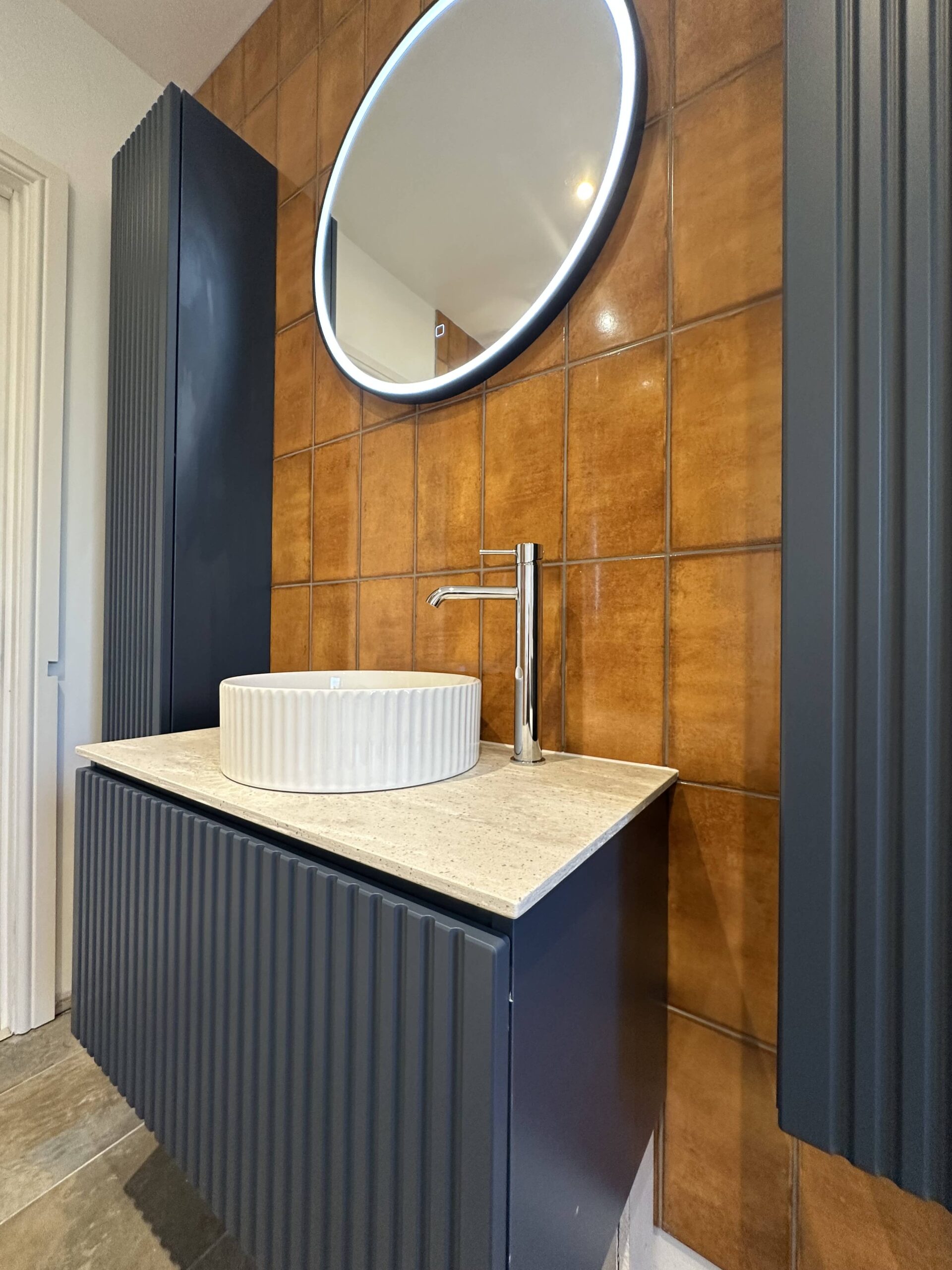 General Plumbing Bathroom (1) Modern bathroom vanity with a round mirror, vessel sink, and brown tiled wall.