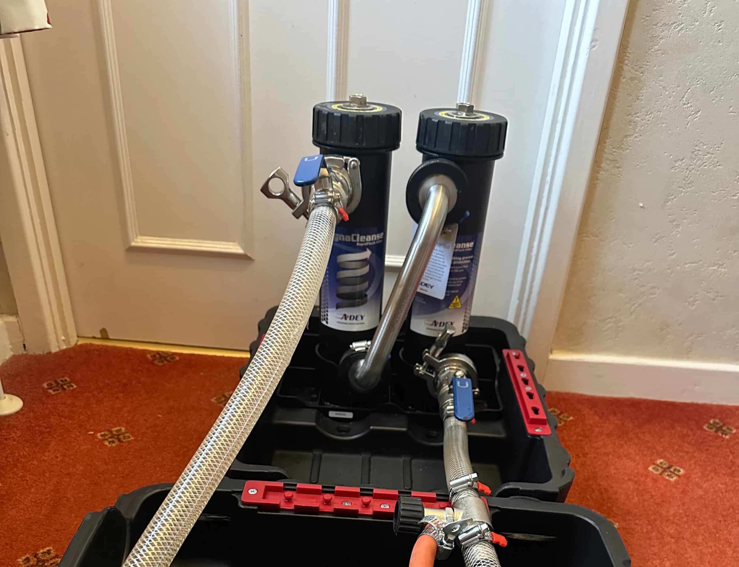 Heat Pumps Two black filter canisters connected by metal hoses inside a plastic container on a red carpet.