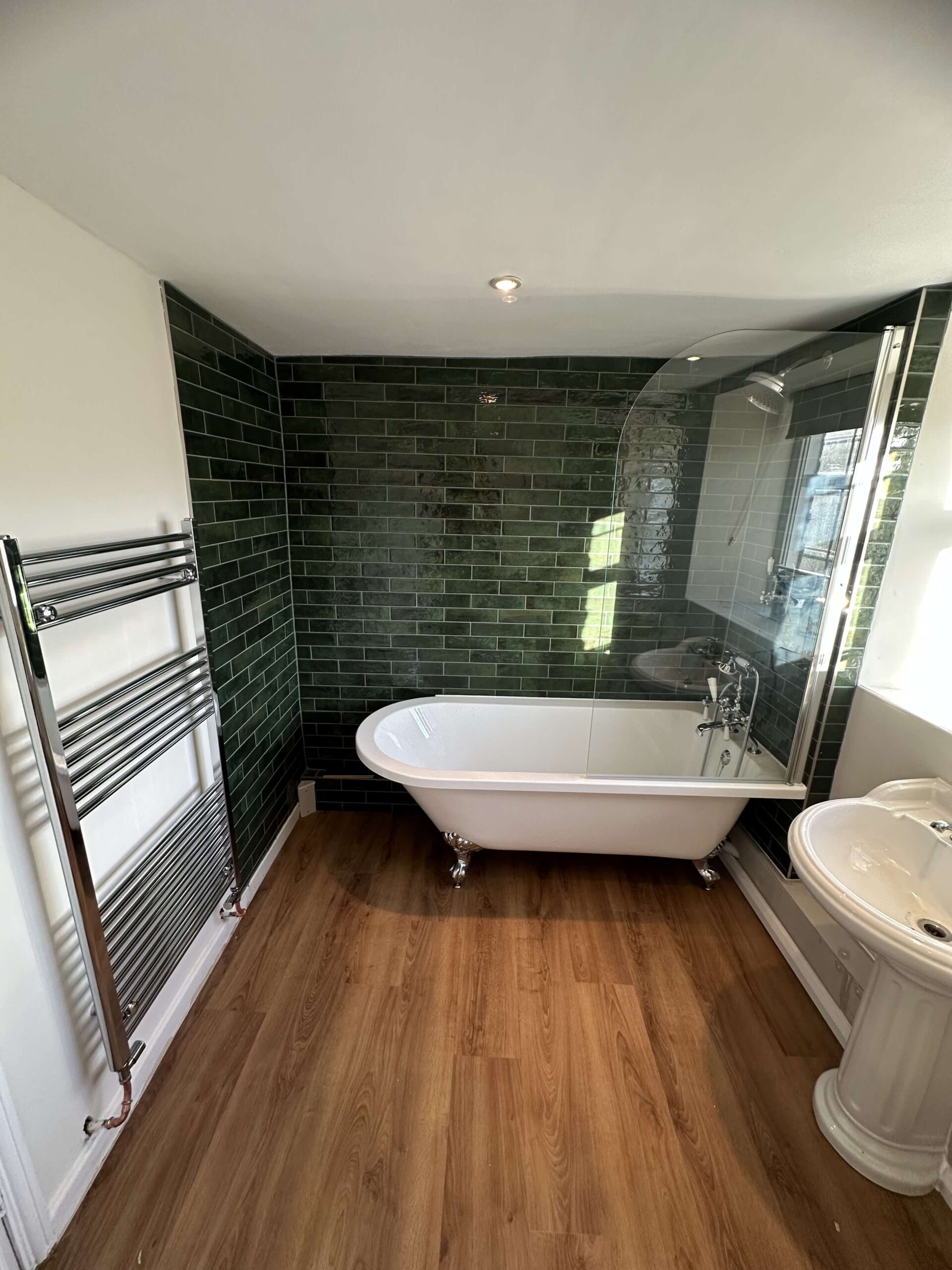 A bathroom with a free-standing bathtub, green tile wall, wooden floor, and a towel rack.