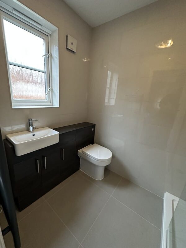 Modern beige tiled bathroom with a window, sink, toilet, and dark cabinet units.