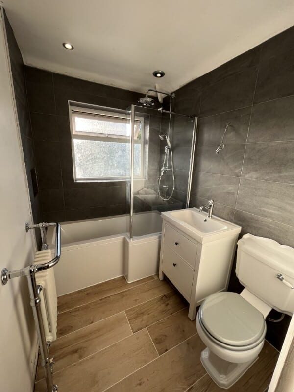 Modern bathroom with black tiled walls, wooden floor, shower, sink, and toilet.