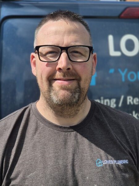 Dan Man with glasses and beard wearing a "LOVES PLUMBING" shirt, standing outdoors in front of a van