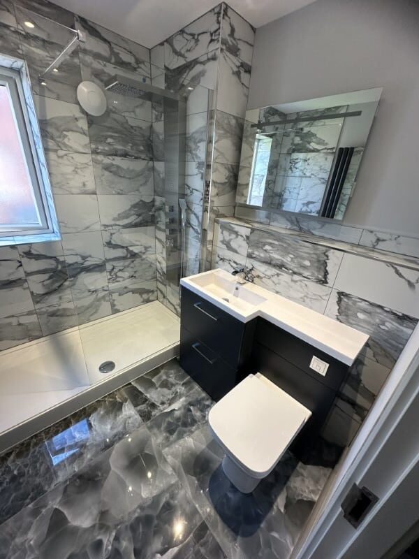 Modern bathroom with grey marble tiles, a walk-in shower, sink, mirror, and toilet.