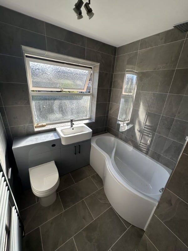Modern bathroom with grey tiles, a white bathtub, sink, toilet, and a frosted window.