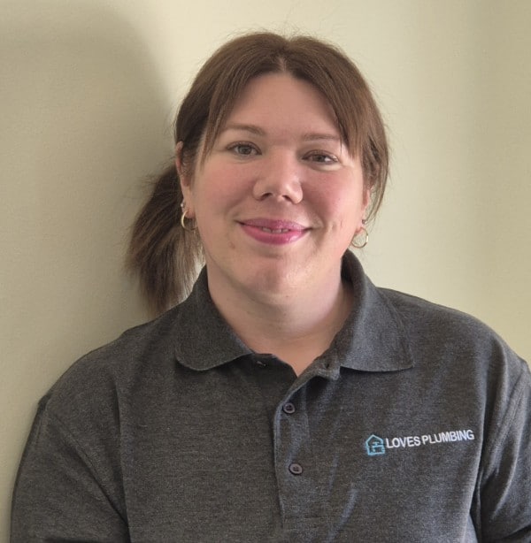 Mel A woman smiling in a grey "Loves Plumbing" shirt, standing against a light-coloured wall.
