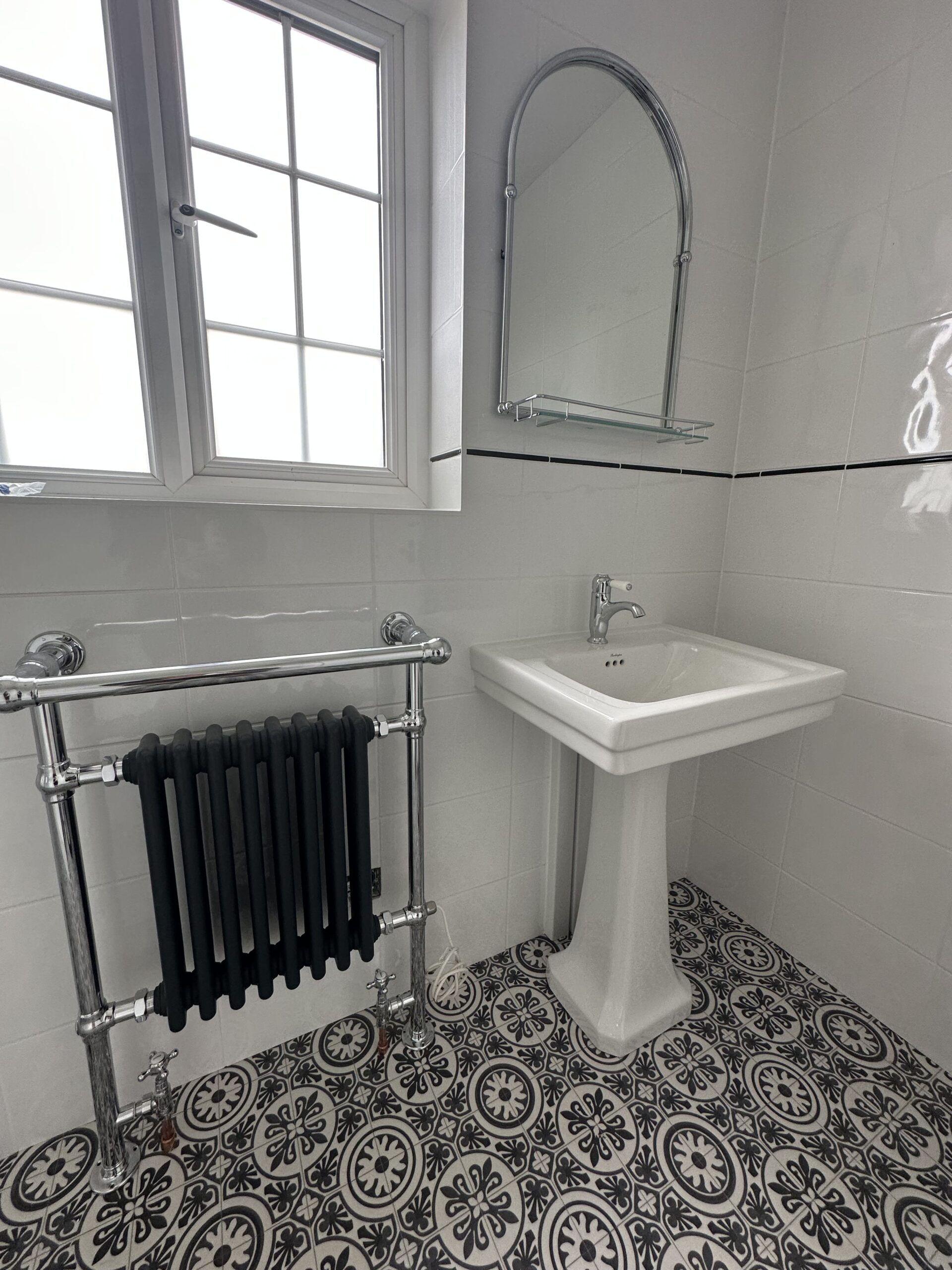 PatternTiles2 Small bathroom with patterned tile floor, white pedestal sink, mirror, radiator, and window.