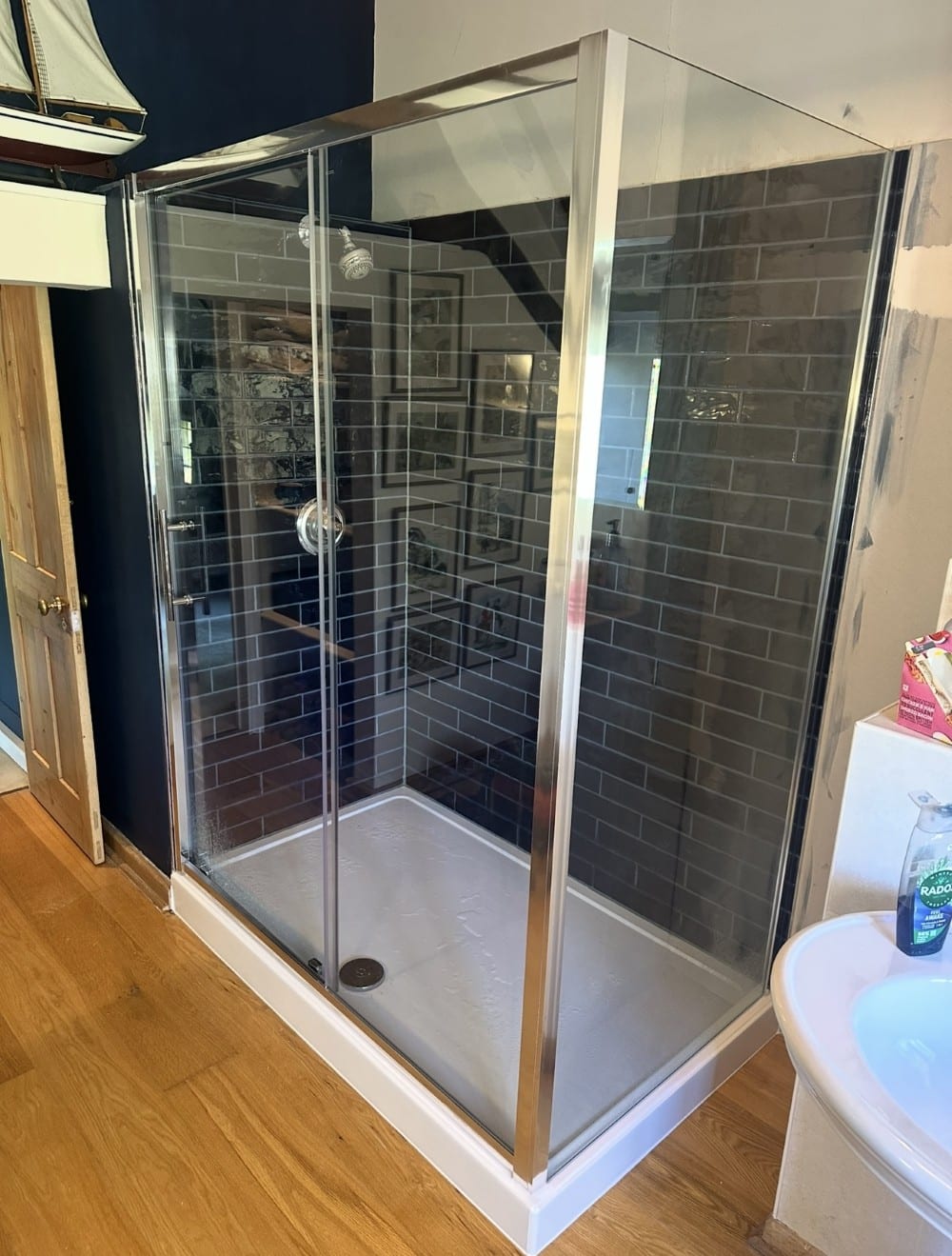 Screenshot Glass shower with black subway tiles and chrome frame, next to wooden floor and white sink.