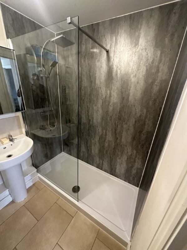 Modern walk-in shower with glass panel, grey walls, and a white pedestal sink nearby.